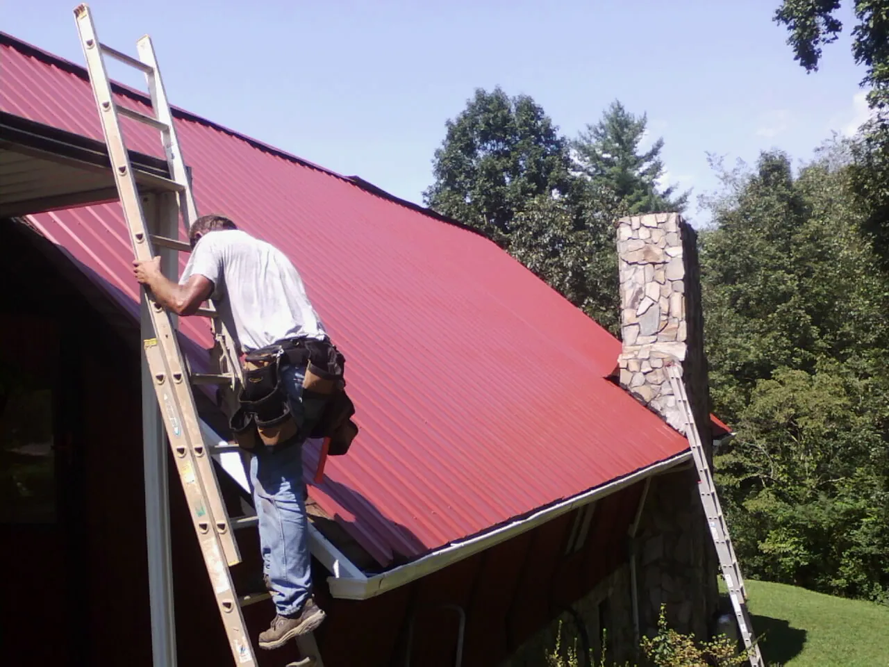 Roofer on a ladder beside a red metal roof and stone chimney — Soffit & Fascia Repair by Roofing Brothers in Moffit